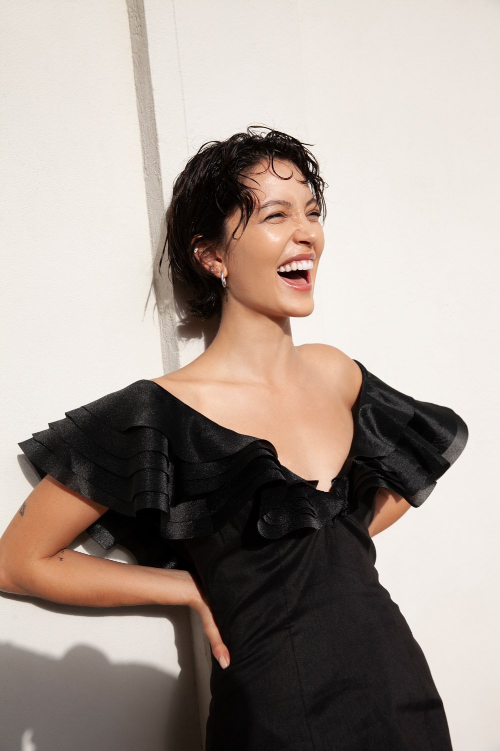 Woman in black dress laughing in front of white wall. Fashion editorial photography with natural light, focus on face, youthful energy.